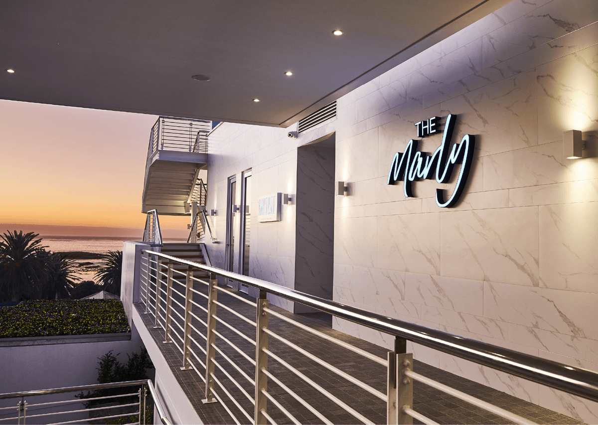 Illuminated "The Marly" sign on the marble entrance facade at dusk, with Baptiste restaurant entrance and ocean sunset views