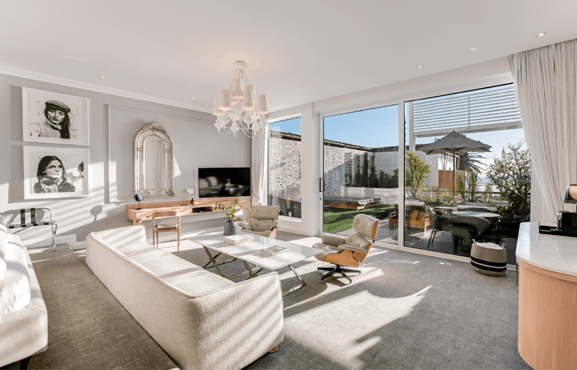 Sunlit luxury suite living area with a cream sofa, Eames lounge chair, marble coffee table, gilt arch mirror, and black-and-white portrait artwork, with large sliding glass doors opening onto a landscaped terrace with sea views.
