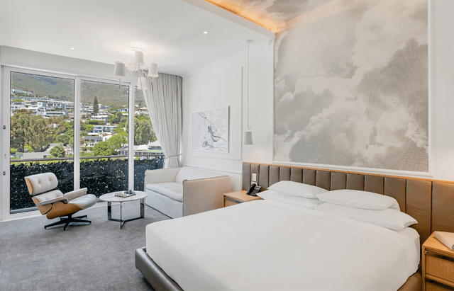 Hotel room with a king bed, cloud-print feature wall, Eames lounge chair, and a compact sofa and side table beside floor-to-ceiling glass doors overlooking a green mountainside and residential Camps Bay.