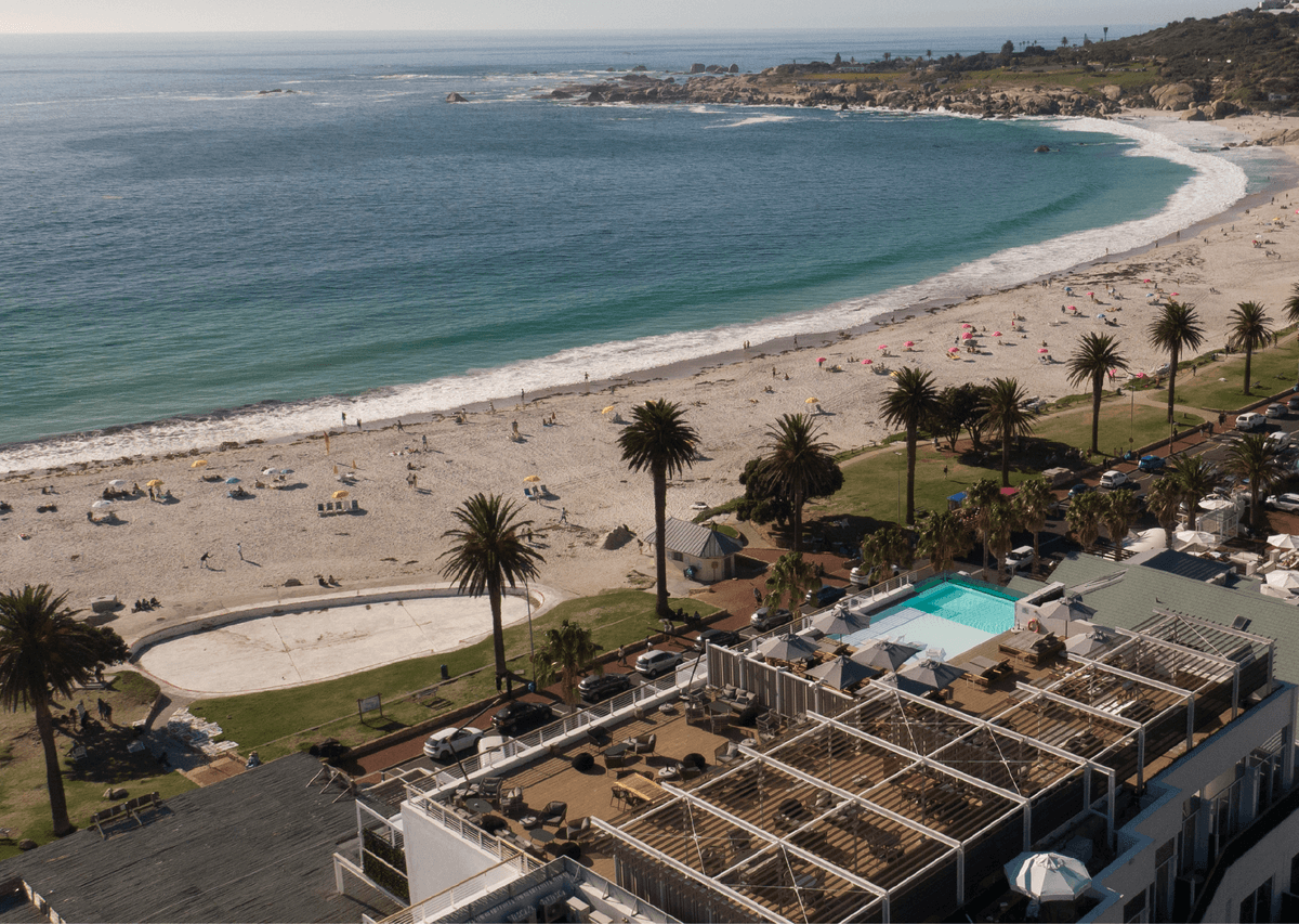 Aerial view of Camps Bay beach with turquoise water, palm-lined white sand, and beachgoers, with The Marly hotel's rooftop terrace and pool visible in the foreground.