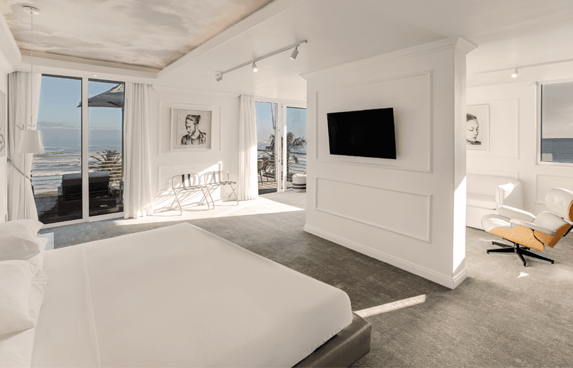 All-white hotel room with a king bed, wall-mounted TV, Eames lounge chair, and black-and-white portrait artworks, with two sets of glass doors opening to sea-view balconies flooded with natural light.