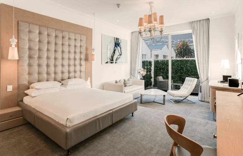 "Hotel room with a king bed beneath a tall tufted upholstered headboard framed in timber, a white sofa, Barcelona-style lounge chair, and marble coffee table, with glass doors opening onto a hedge-screened balcony with mountain views beyond.