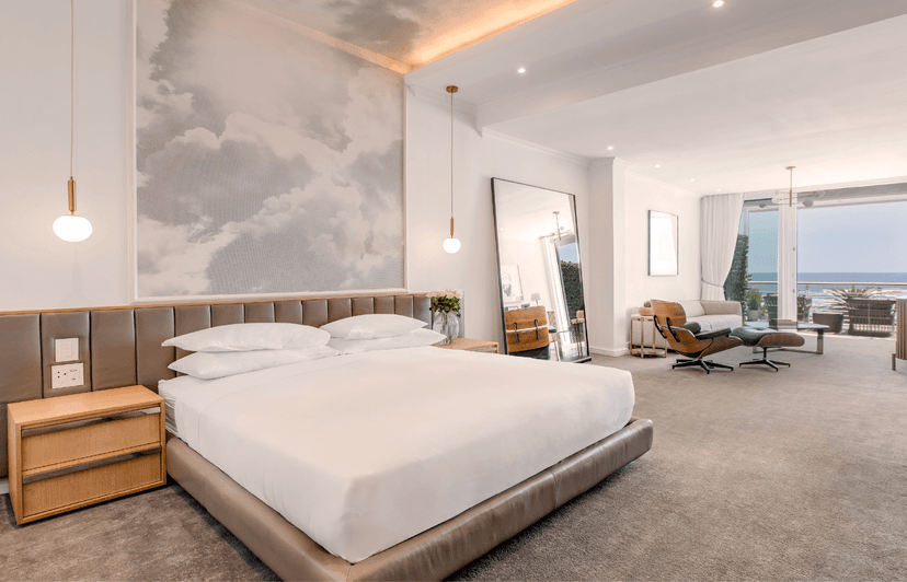 Spacious hotel room with a king bed beneath a cloud-painted feature wall, gold pendant lights, a leaning full-length mirror, Eames lounge chair, and open glass doors leading to a sea-facing terrace.