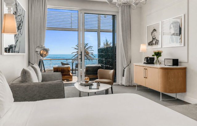 Elegant suite with a king bed, grey sofa, velvet accent chair, and marble coffee table, with a timber sideboard topped with fresh flowers and a Marshall speaker, and glass doors opening to a sea-view balcony with palm trees.