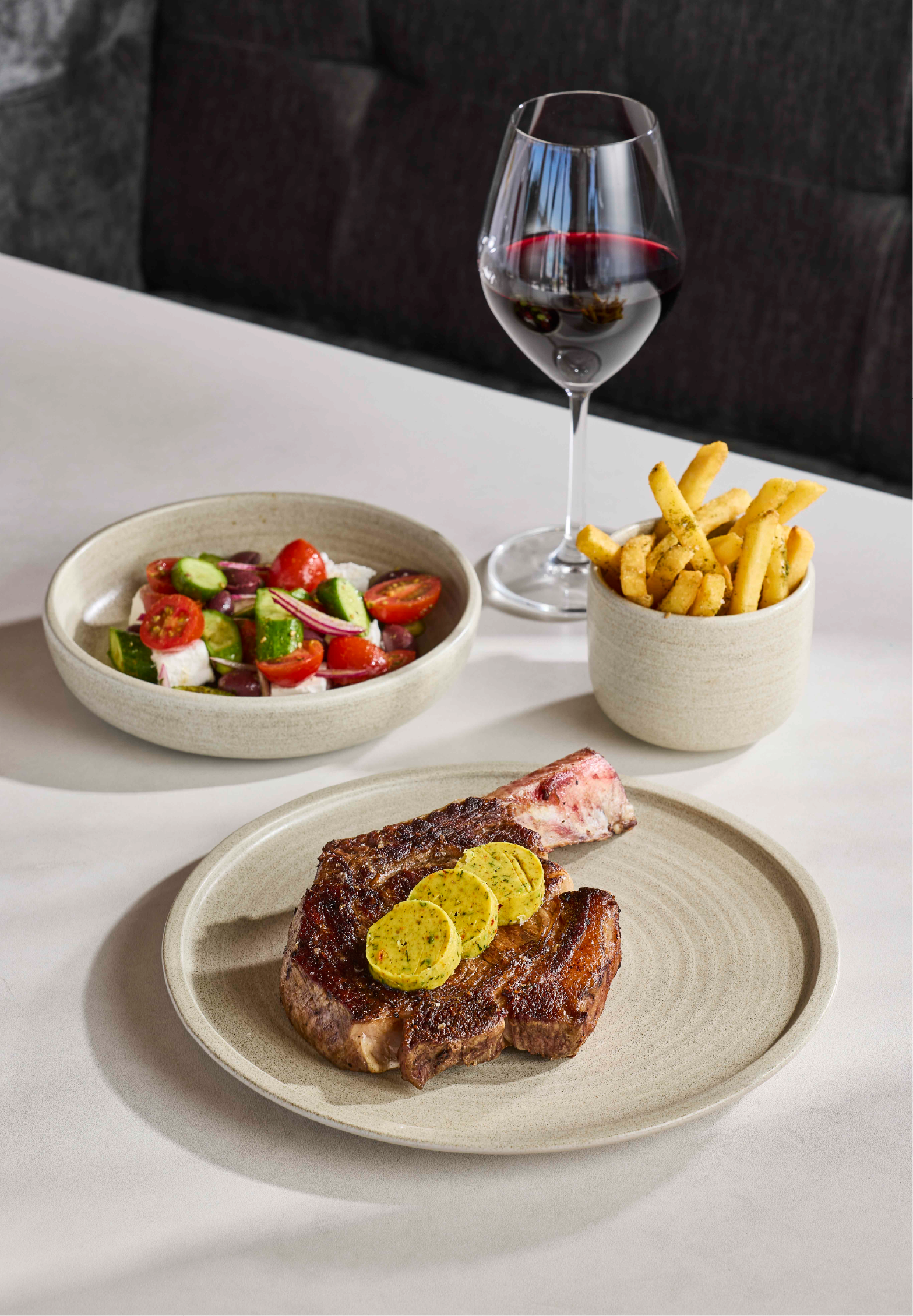 A bone-in ribeye steak topped with herb butter discs, served alongside a Greek salad, a cup of seasoned fries, and a glass of red wine on a white table.