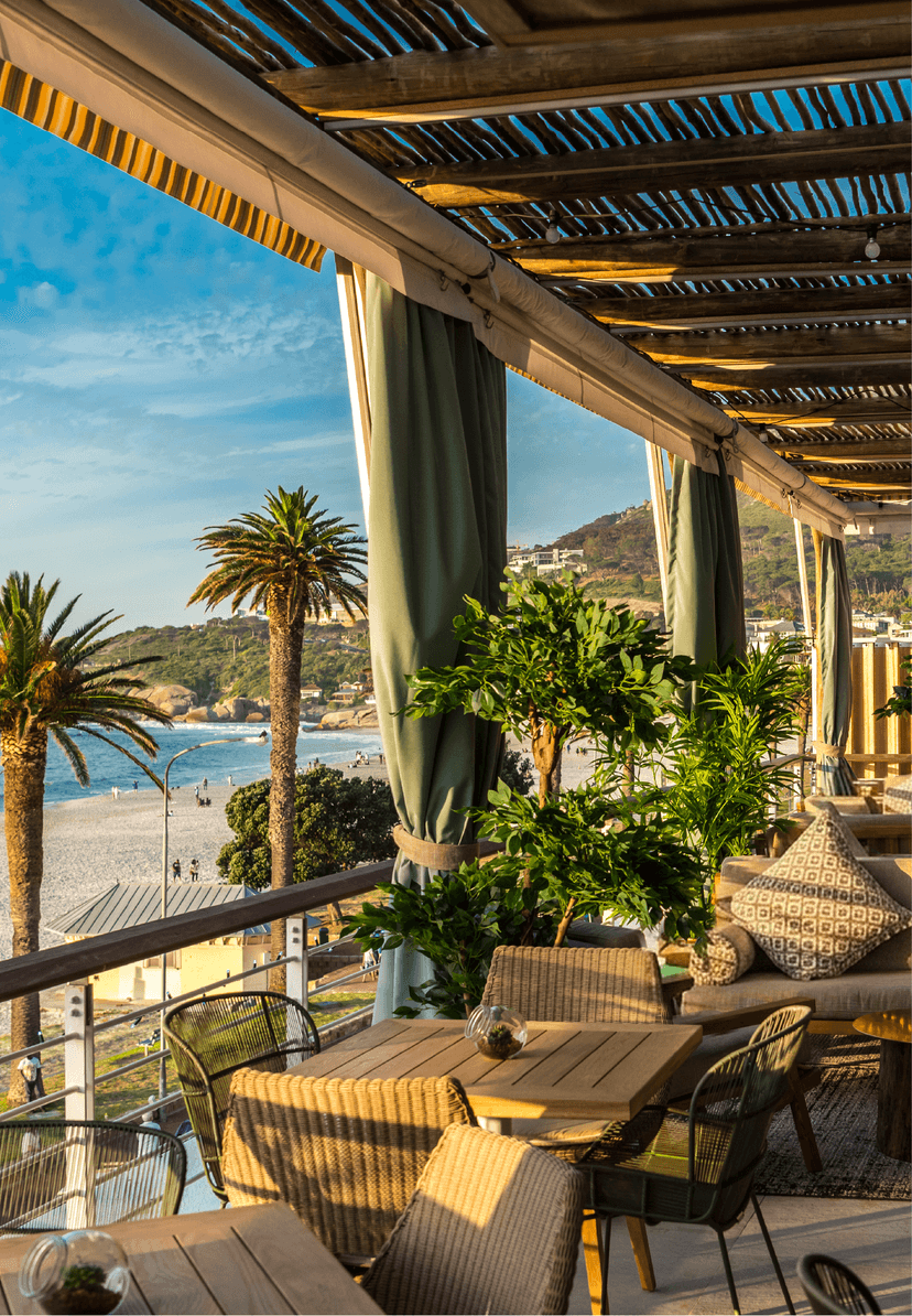 Chinchilla's open-sided terrace at golden hour, with cushioned wicker lounge seating, lush potted greenery, green drape curtains, and a direct view over Camps Bay beach, palm trees, and the Atlantic.