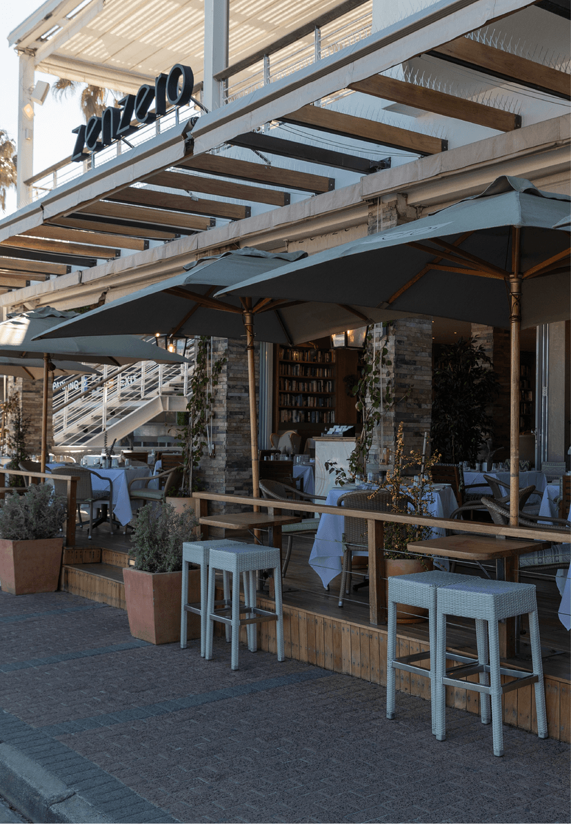 Zenzero restaurant's street-facing exterior at dusk, with sage-green umbrellas, white-clothed tables, bar-height stools, terracotta herb planters, and the Zenzero sign above the entrance.
