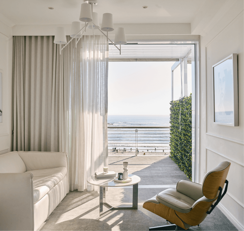 Bright hotel room sitting area with a white sofa, Eames lounge chair, and marble side table, with sheer curtains framing an open glass door leading to a timber deck balcony with beach and ocean views beyond.