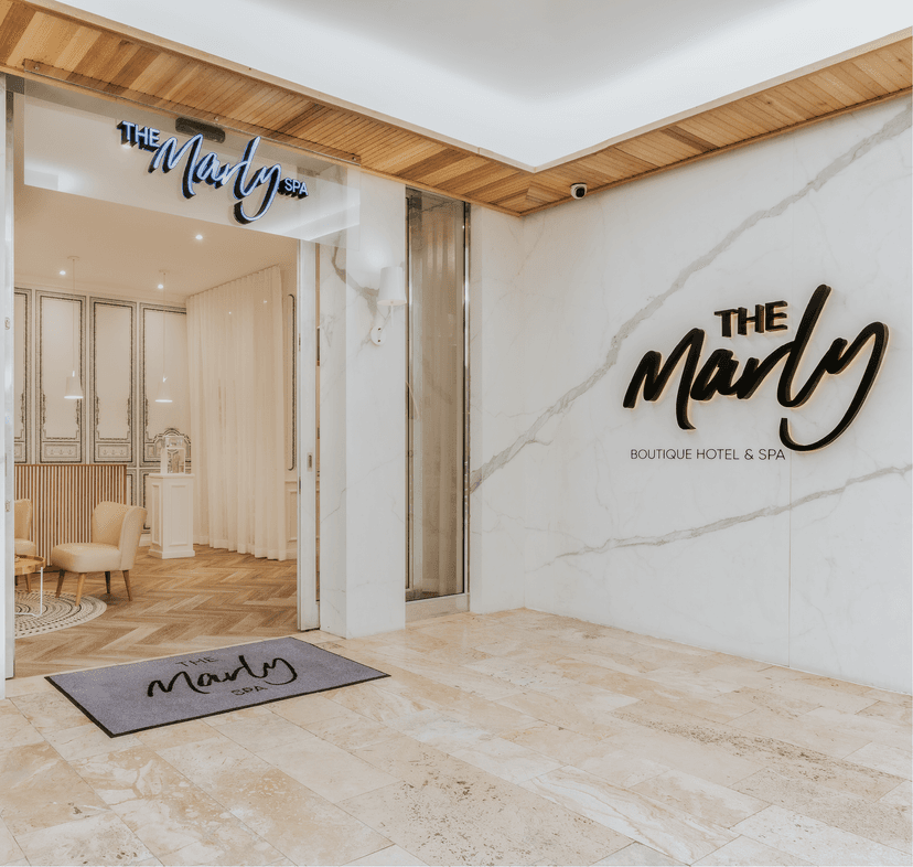 The entrance to The Marly Spa, with a neon spa sign above the open glass door, a branded welcome mat on travertine flooring, and 'The Marly Boutique Hotel & Spa' lettering mounted on a marble feature wall.