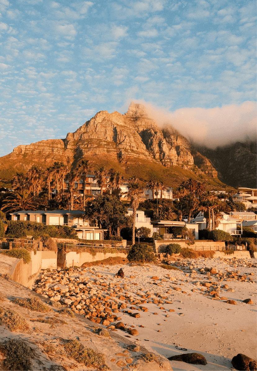 Glen beach at golden hour, with rounded pebbles in the foreground, palm-lined residential properties in the middle ground, and the cloud-draped Twelve Apostles glowing amber in the late afternoon sun.