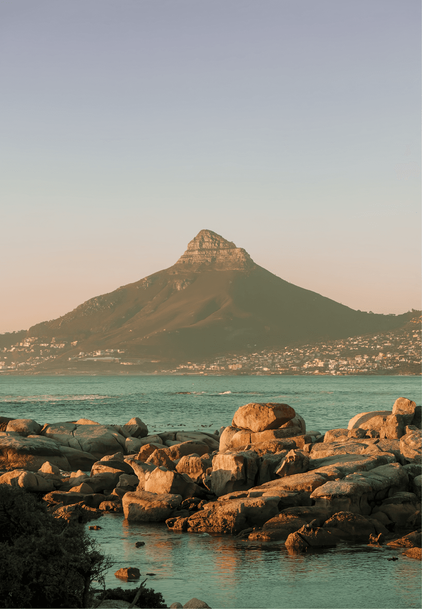 Warm dusk light on the granite boulder shoreline looking across the bay toward Lion's Head, with turquoise water and the city of Cape Town spread across the hillside below.