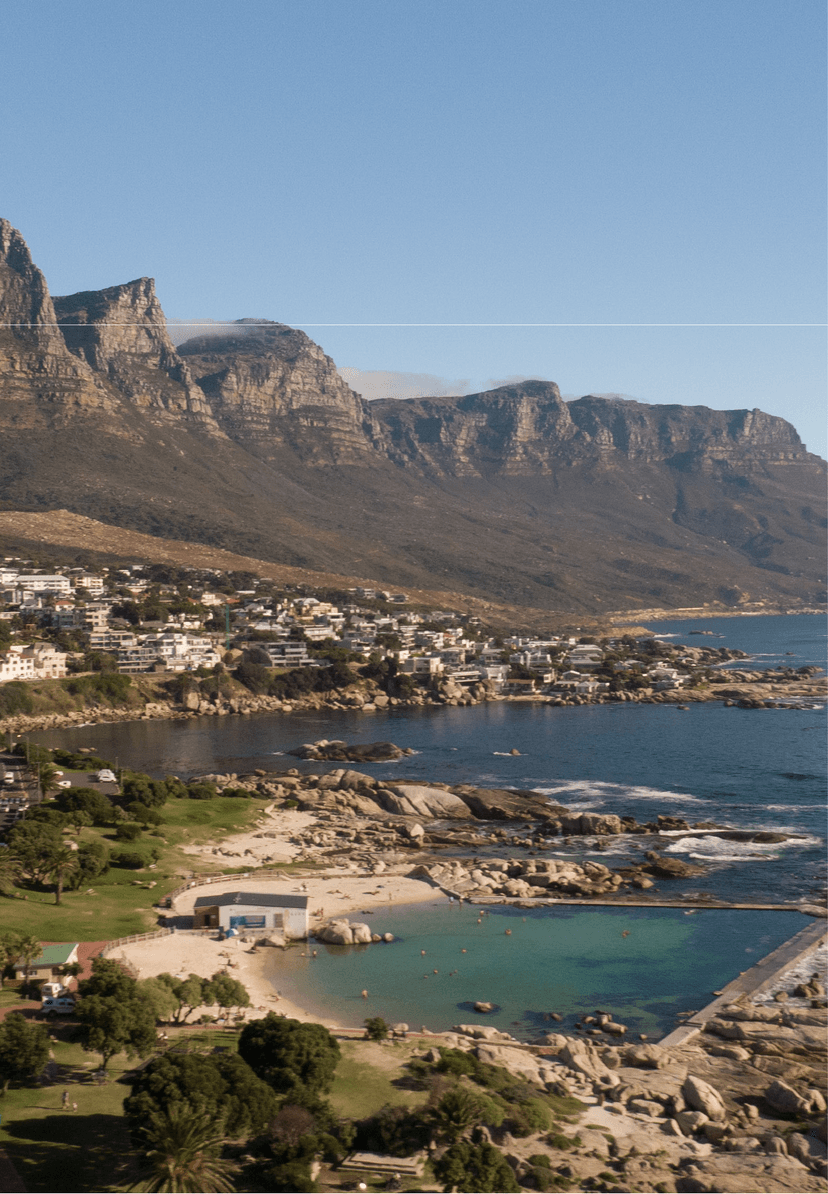 Elevated view of the Camps Bay coastline on a clear day, showing the tidal pool and main beach below, rocky outcrops along the shore, and the Twelve Apostles stretching along the horizon.
