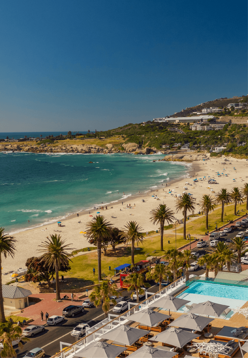 Aerial view of Camps Bay beach on a busy summer day, with turquoise water, a packed white sand beach lined with palm trees, and The Marly's rooftop pool visible in the foreground.