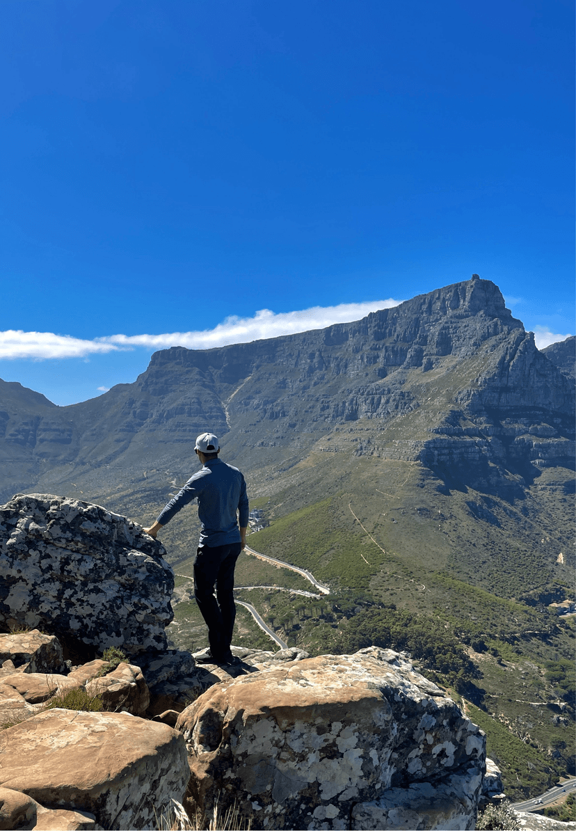 A hiker in a cap and blue top stands on a rocky ledge looking up toward Table Mountain, with a winding mountain road and lush green valley visible below under a vivid blue sky.