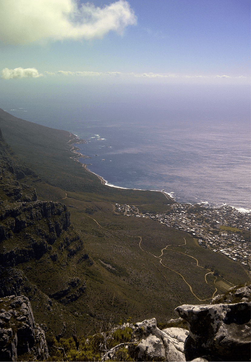 Dramatic bird's-eye view from the top of Table Mountain looking south along the Atlantic coastline, with hiking trails cutting through the fynbos, a small coastal town far below, and the vast ocean beyond.