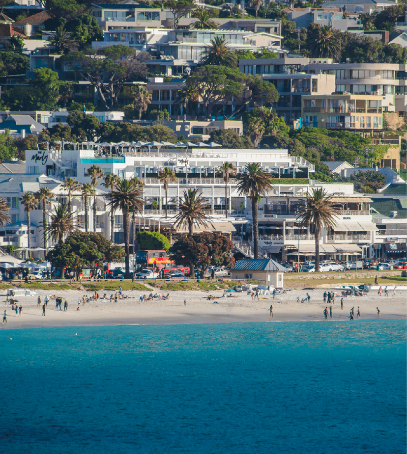 View from the water looking toward Camps Bay beachfront, with The Marly hotel's white façade, rooftop pool and terraces clearly visible above the palm-lined strip, surrounded by lush hillside residential properties.