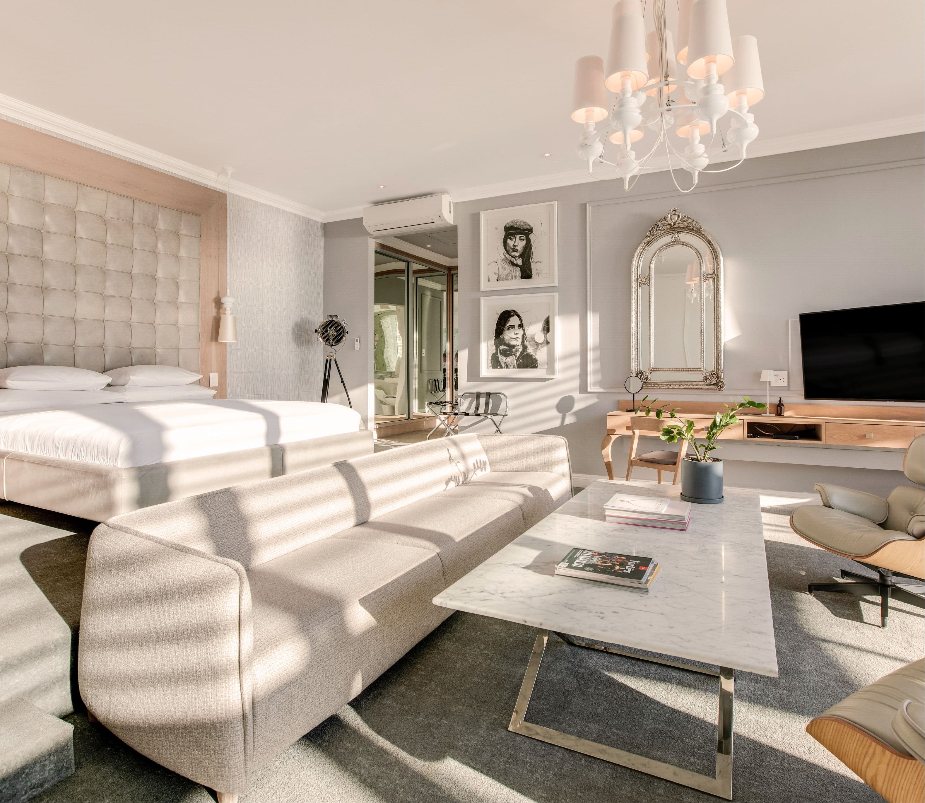 Bright hotel suite with a tufted upholstered king bed, cream sofa, marble coffee table, and Eames lounge chair, decorated with black-and-white portrait artworks, a gilt arch mirror, and a white chandelier above.