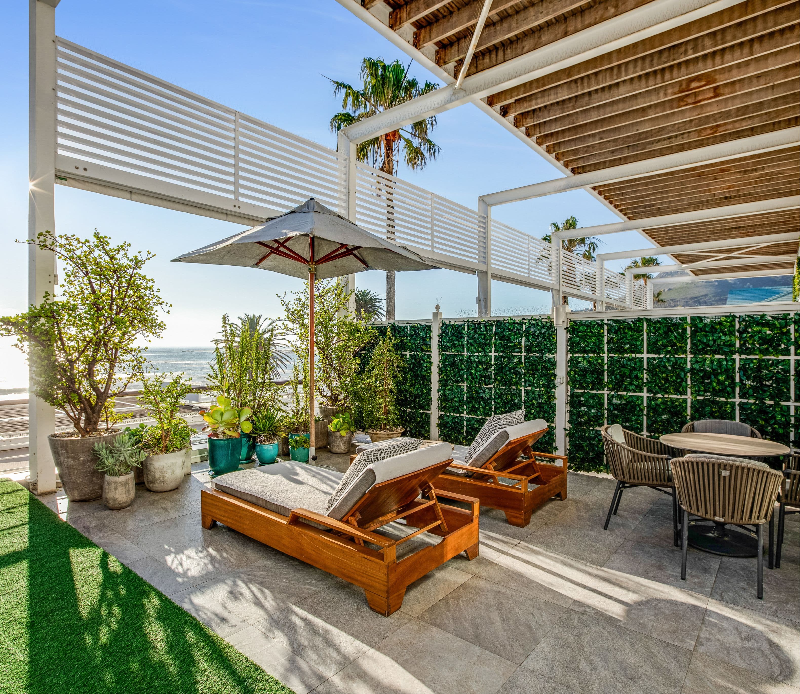 Sunny private terrace with two cushioned teak sun loungers and an umbrella, surrounded by abundant potted plants and a green hedge screen, with palm trees and the ocean visible in the background.