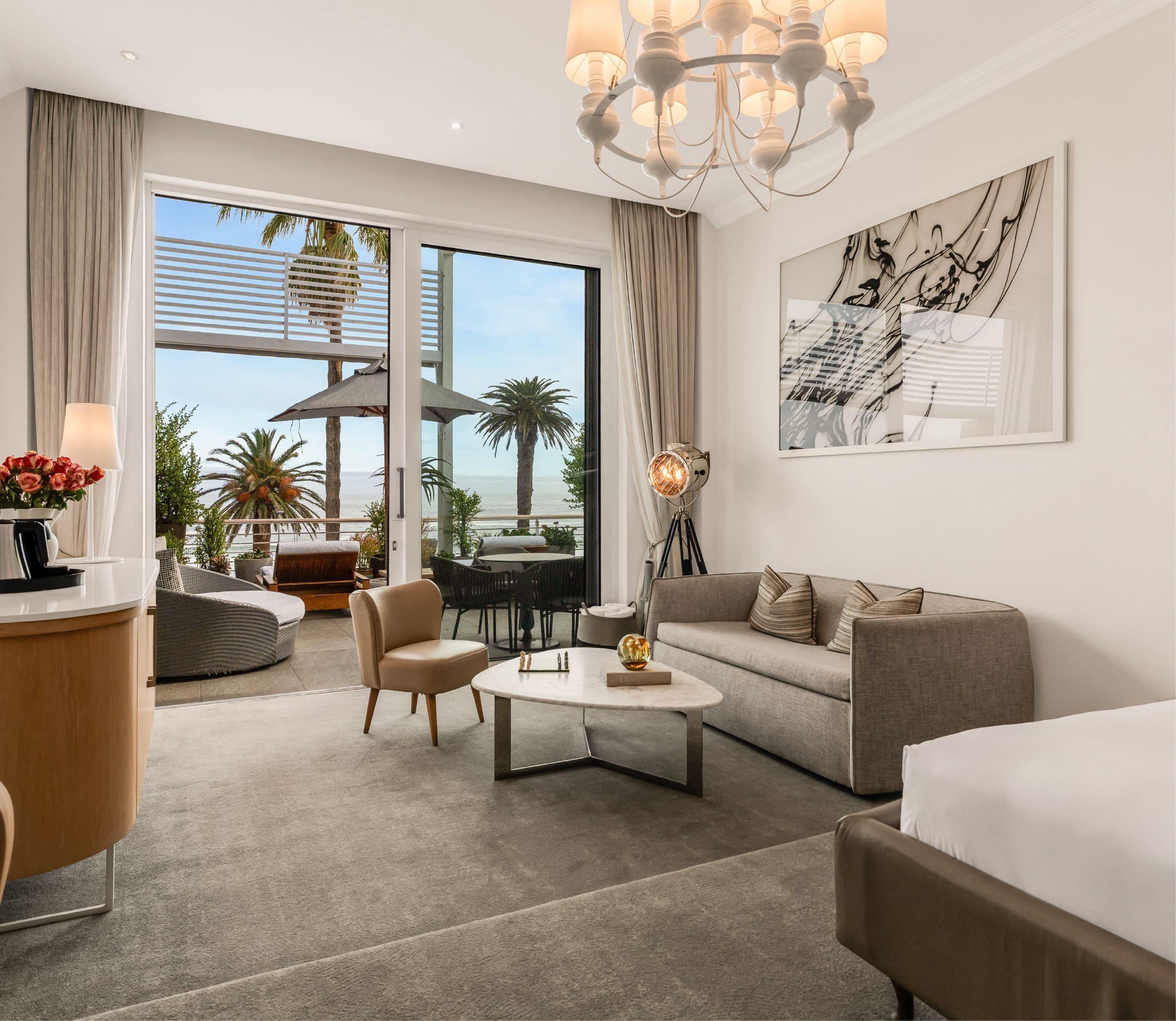 Elegant suite living area with a grey sofa, velvet accent chair, and marble coffee table beneath a white chandelier, with large glass doors opening onto a plant-lined terrace overlooking the ocean.