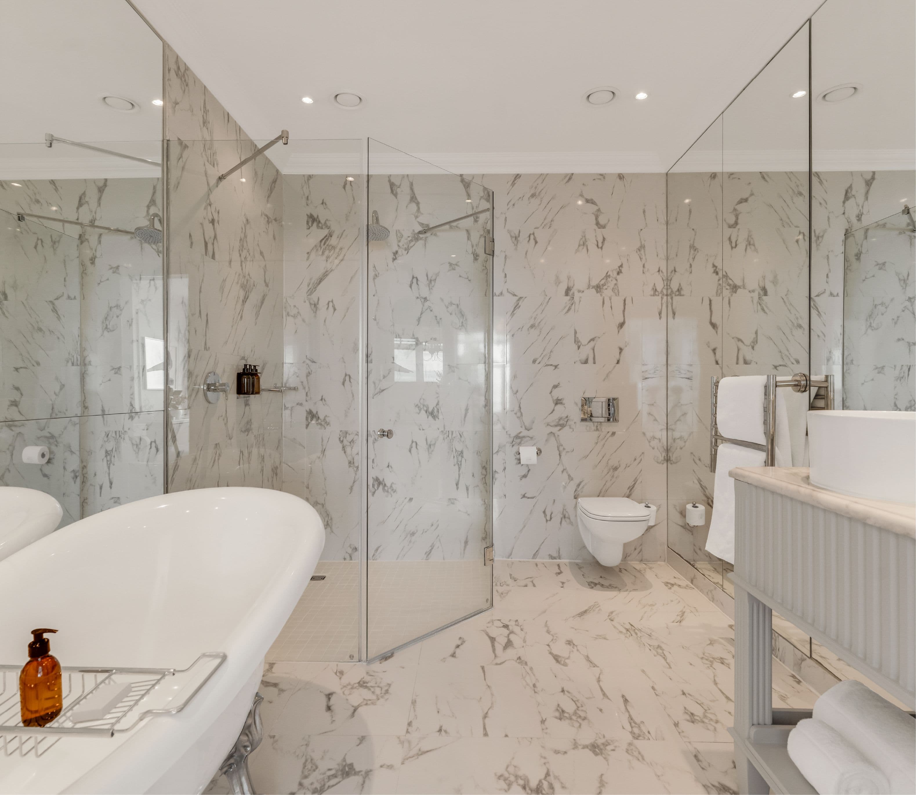Spacious luxury en-suite bathroom clad in white and grey marble, featuring a freestanding soaking tub, glass-enclosed walk-in shower, wall-mounted toilet, and double vanity with illuminated mirrors.