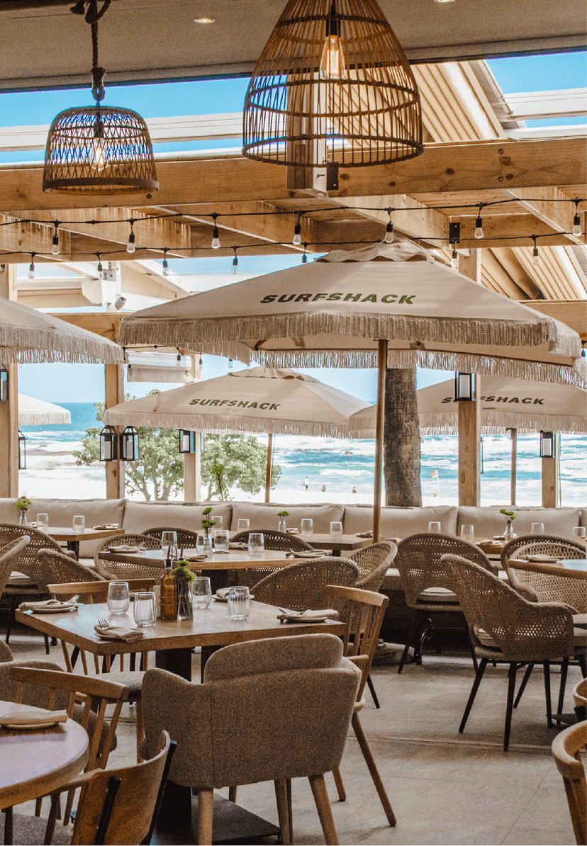 Surfshack restaurant's airy interior with woven rattan pendant lights, fringed branded umbrellas, and a mix of upholstered and woven chairs, looking out over the beach and breaking waves.