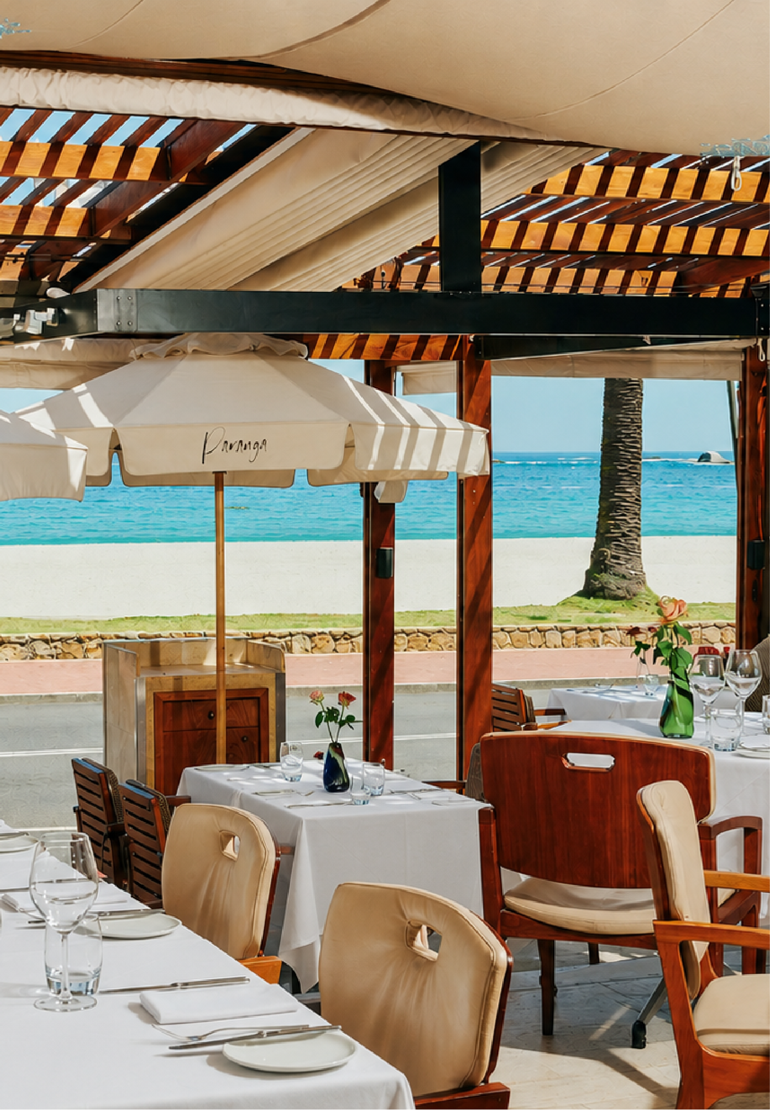 Paranga restaurant's open terrace set for lunch, with white-clothed tables, a branded striped umbrella, teak chairs, and a direct view of Camps Bay beach, palm trees, and the ocean.