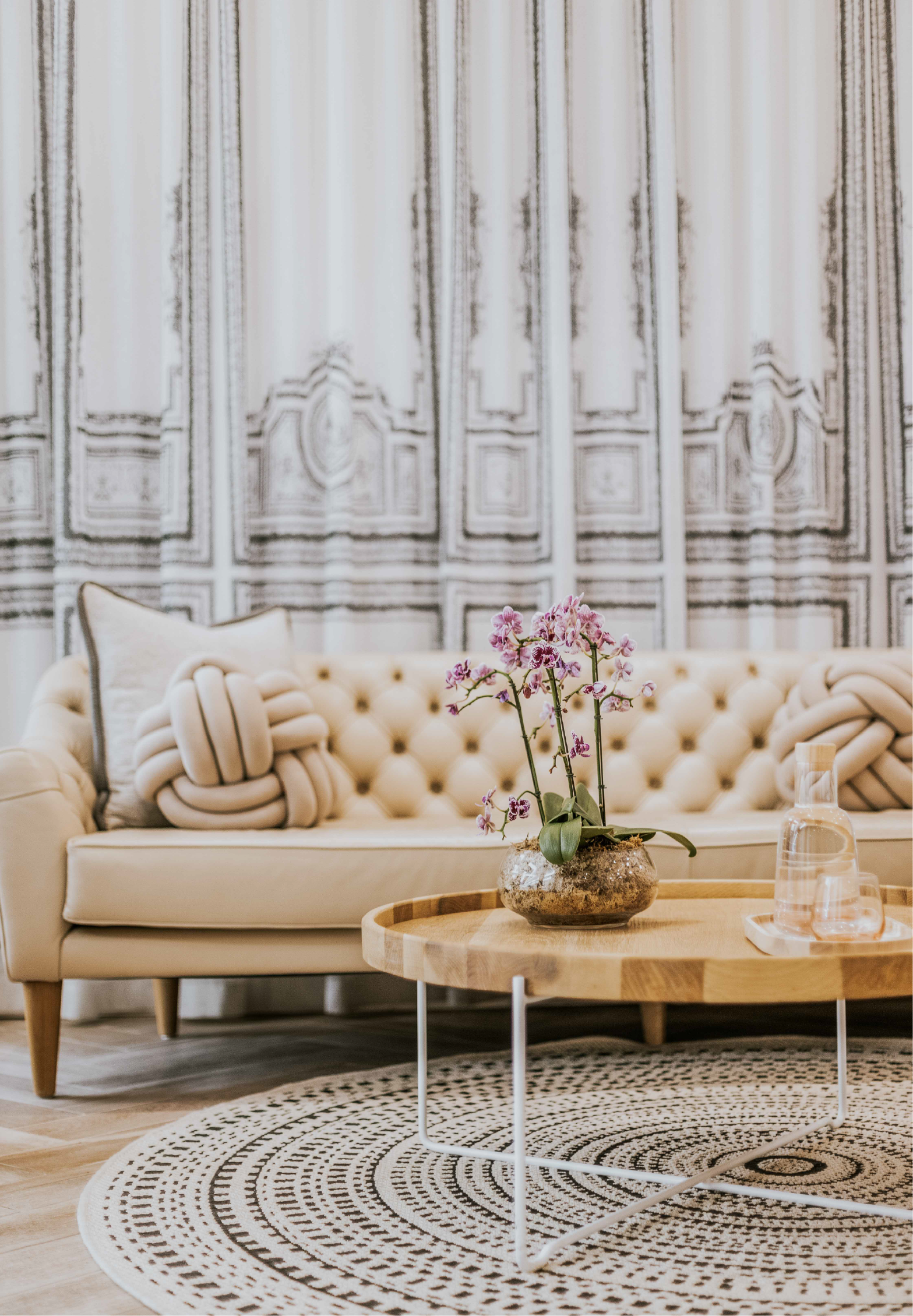 The Marly Spa's elegant waiting lounge, featuring a cream button-tufted sofa with knotted cushions, a round wood and acrylic coffee table with a potted orchid, and ornate architectural wallpaper behind.