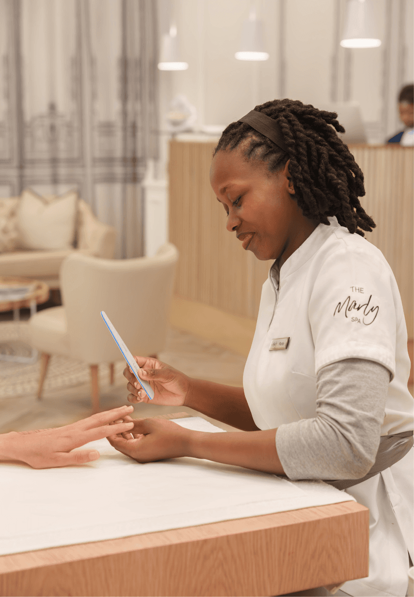 A The Marly Spa therapist in a white uniform performs a manicure treatment, carefully filing a guest's nails at a light timber treatment station in the spa's softly lit interior.