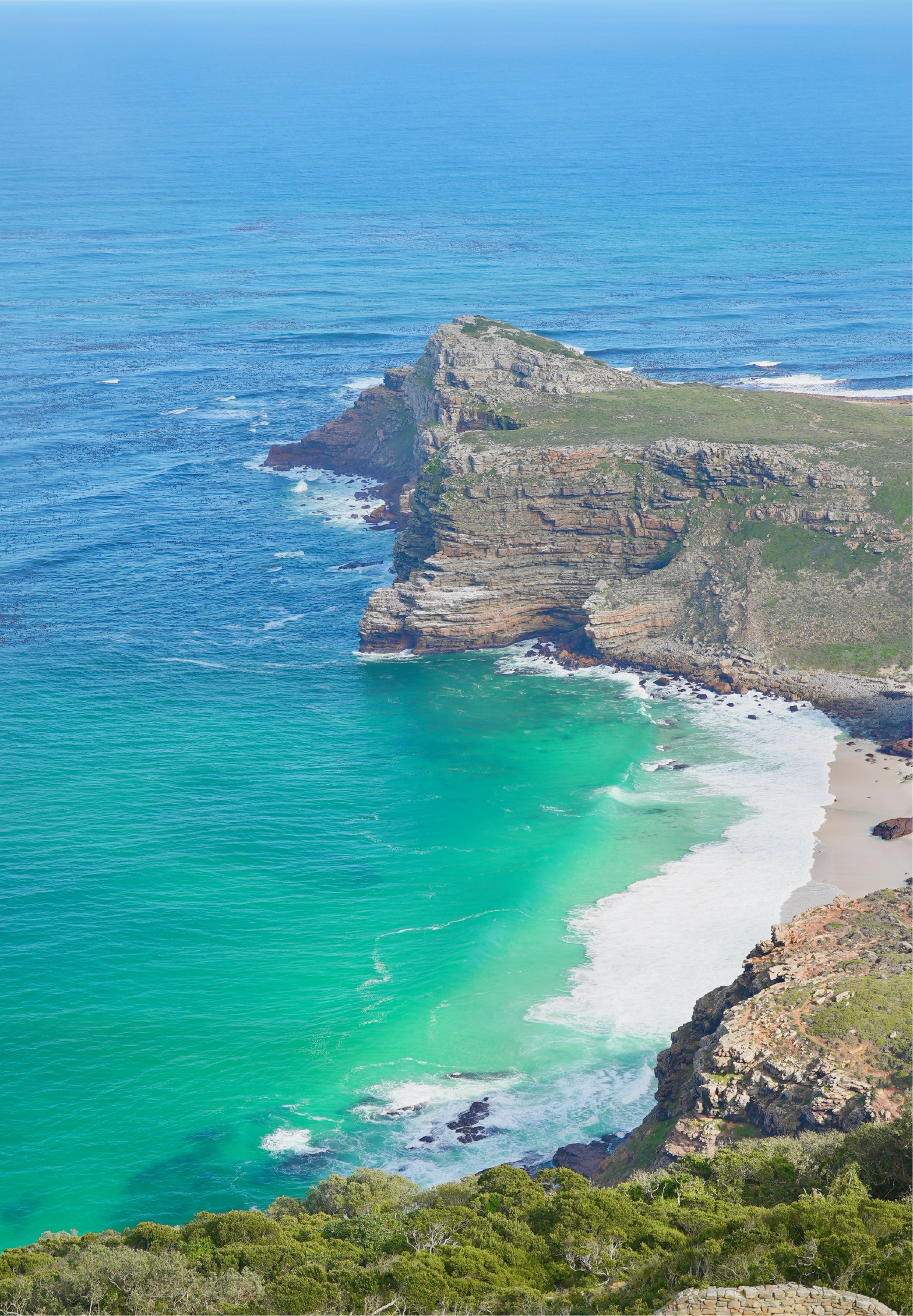 Aerial view of Cape Point's dramatic layered sandstone headland jutting into the deep blue Atlantic Ocean, with a white sandy beach and vivid turquoise water in the sheltered cove below.