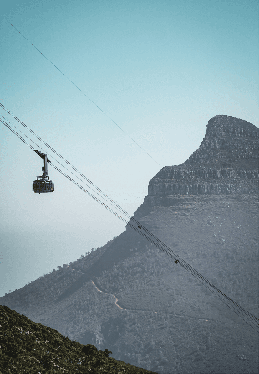 The red Table Mountain Aerial Cableway car ascending its cable against a pale blue sky, with Lion's Head and the mountain's lower slopes visible below.