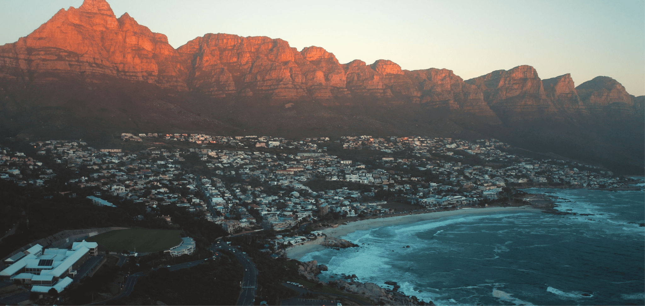Aerial view of Camps Bay at dusk, with the Twelve Apostles mountain range glowing amber in the last light of sunset, the curved white sand beach, and residential neighbourhood spread below.