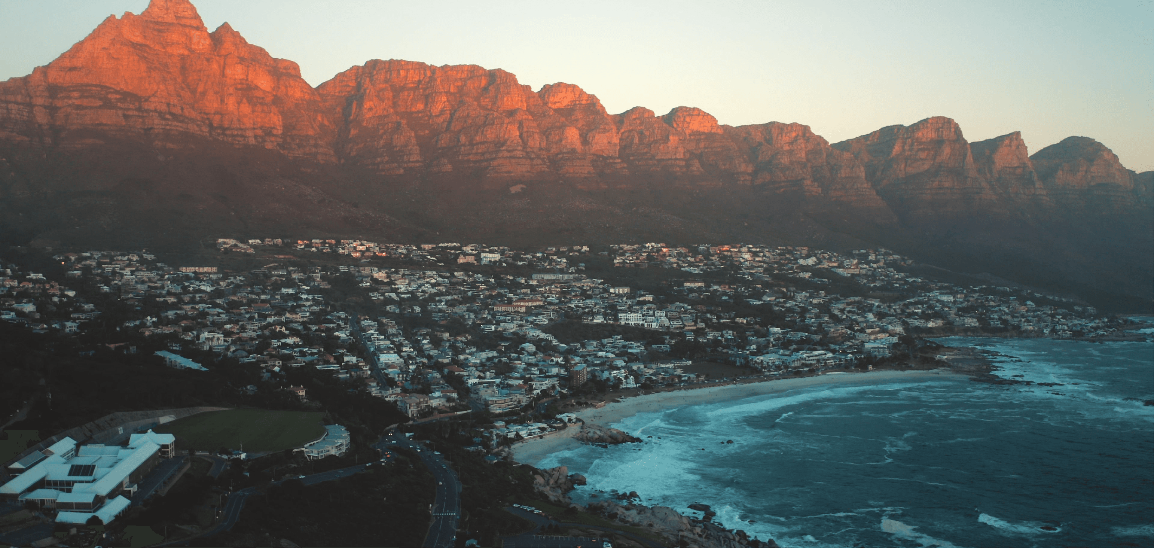 Aerial view of Camps Bay at dusk, with the Twelve Apostles mountain range glowing amber in the last light of sunset, the curved white sand beach, and residential neighbourhood spread below.