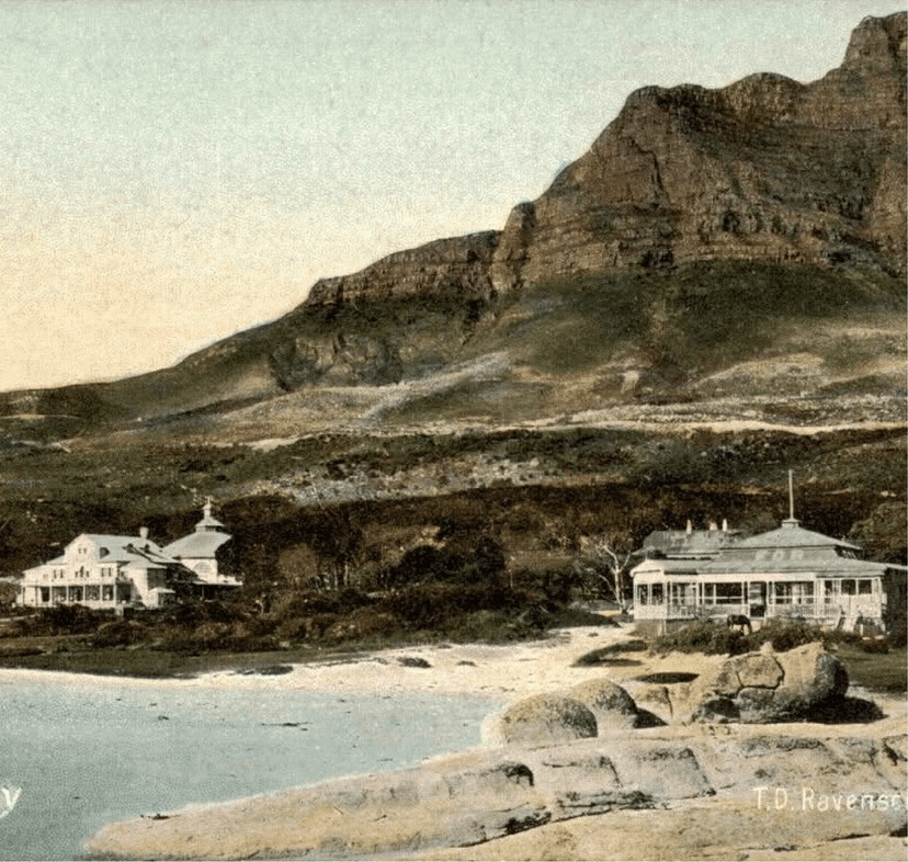 Hand-tinted early 1900s postcard of Camps Bay showing two colonial-era buildings beside the beach, backed by a dramatic rocky mountain slope, credited to T.D. Ravenscroft.