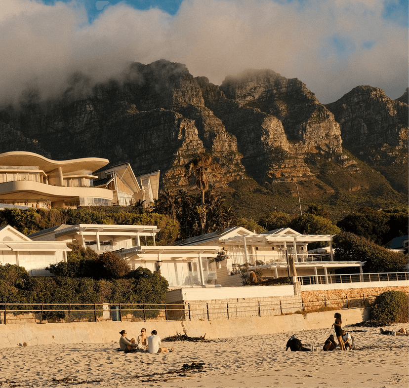 People relaxing on Glen Beach at golden hour, with low-slung modern homes and lush greenery in the middle ground and the cloud-draped Twelve Apostles rising dramatically behind.