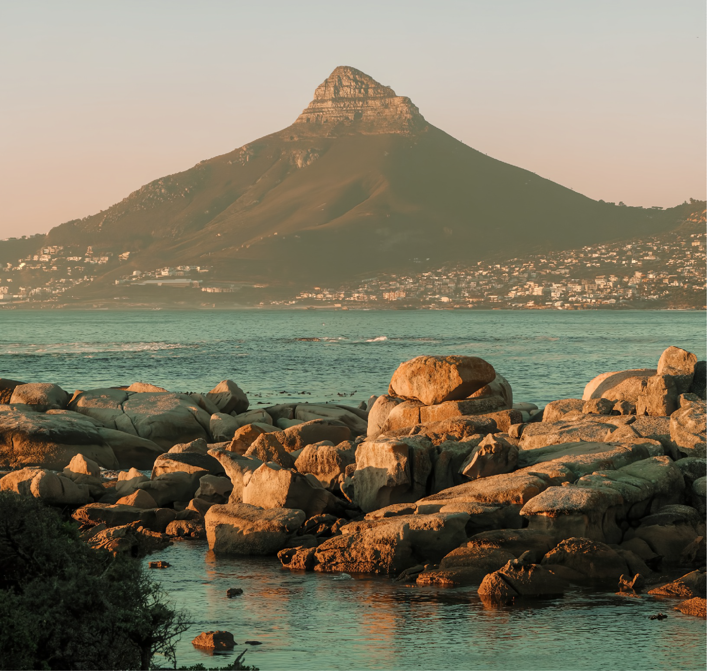 Warm late-afternoon light on the granite boulder shoreline at Bakoven, with turquoise water in the foreground and Lion's Head rising behind the city across the bay.