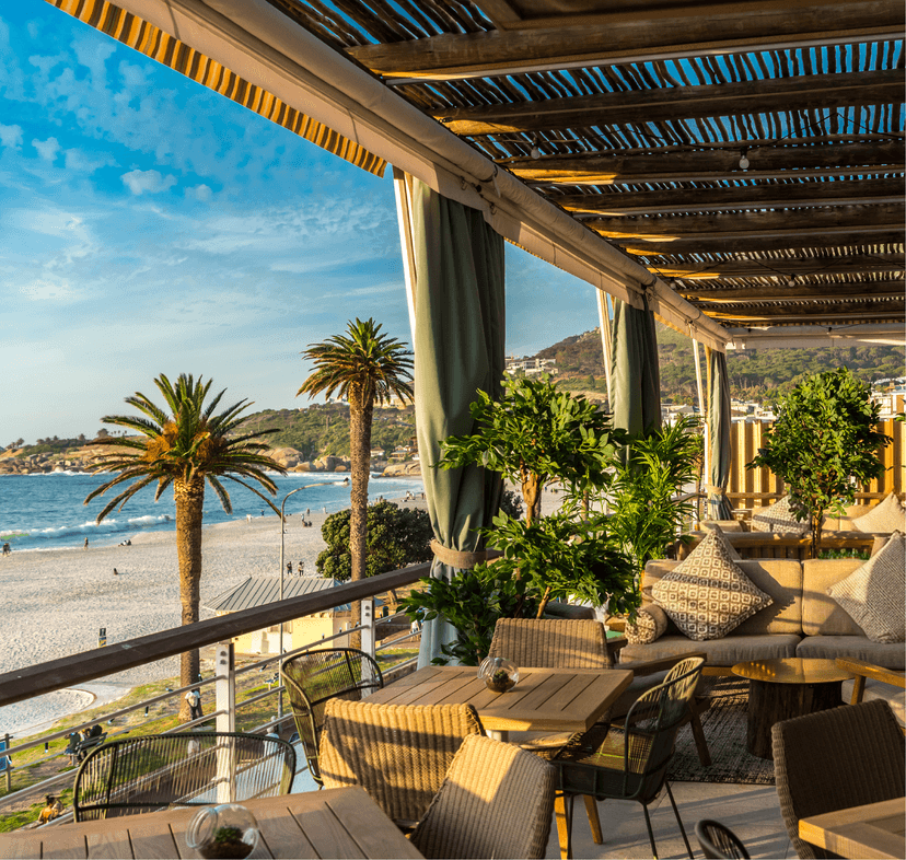 Chinchilla's open-sided rooftop terrace at golden hour, with cushioned wicker lounge seating, lush potted greenery, green drape curtains, and a sweeping view of Camps Bay beach and the Atlantic.