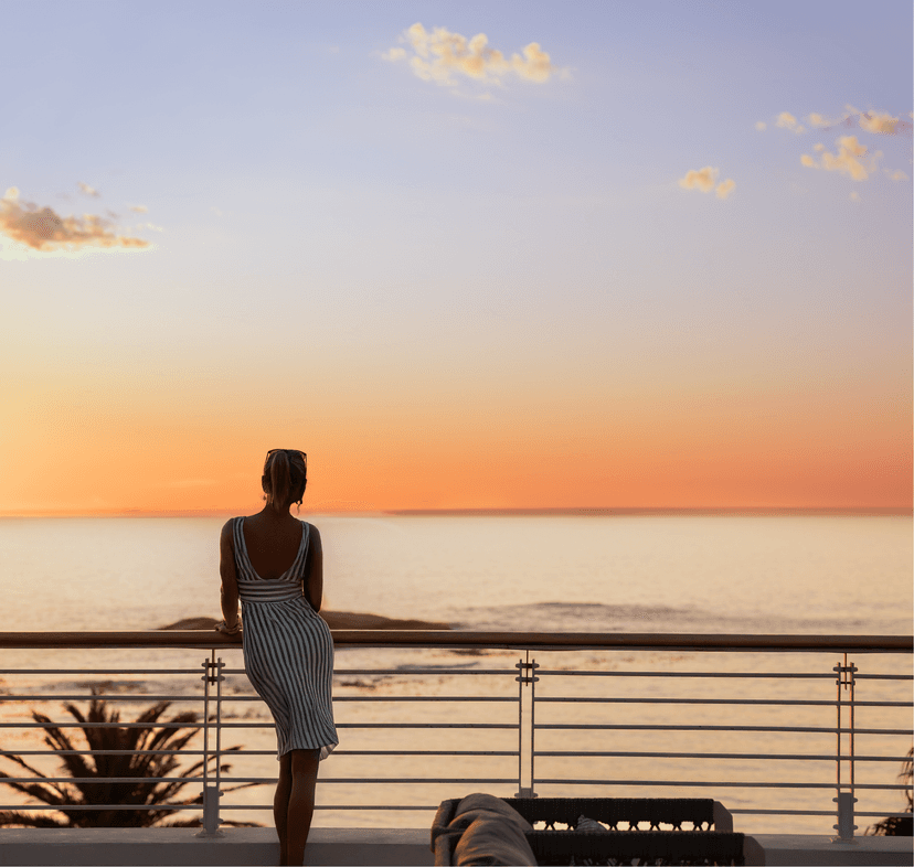 A woman in a striped backless dress stands at a railing, silhouetted against a vivid orange and peach sunset over a calm ocean.