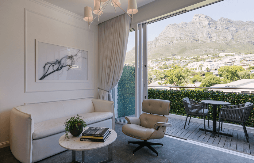Bright hotel room sitting area with a white leather sofa, Eames lounge chair, and marble side table, with open sliding doors leading to a balcony with outdoor dining furniture overlooking the Twelve Apostles mountain range.