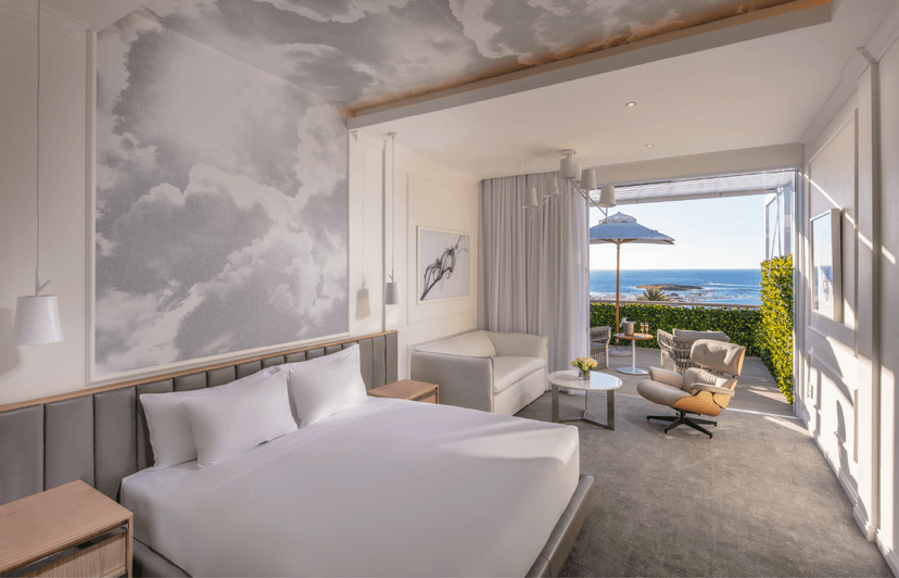 Airy hotel room with a king bed, cloud-painted feature wall and ceiling, cream sofa, and Eames lounge chair, with open glass doors revealing a sea-view balcony with an umbrella and woven chairs.