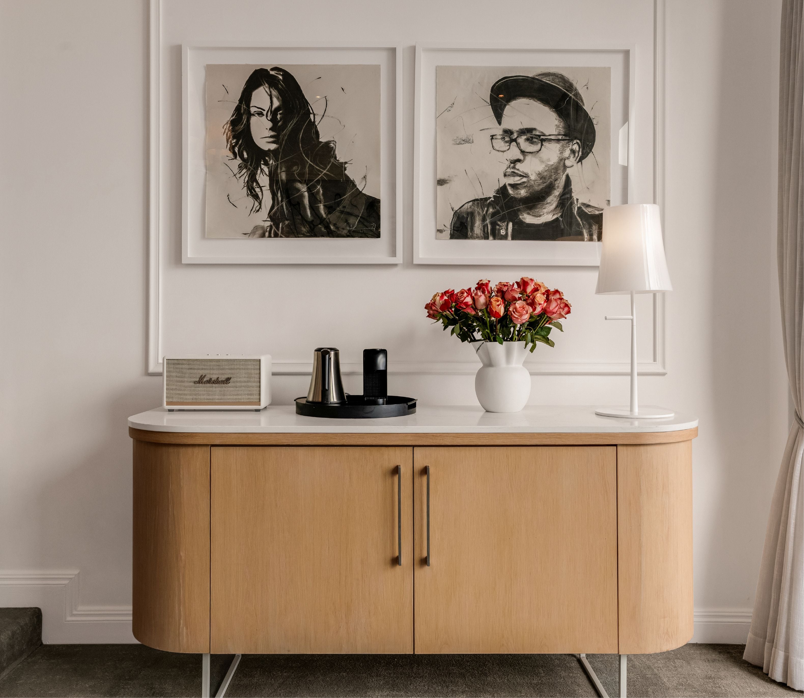 Charcoal portrait artworks above a wooden credenza with Marshall speaker, Nespresso machine and fresh roses in a Deluxe Sea-Facing Suite