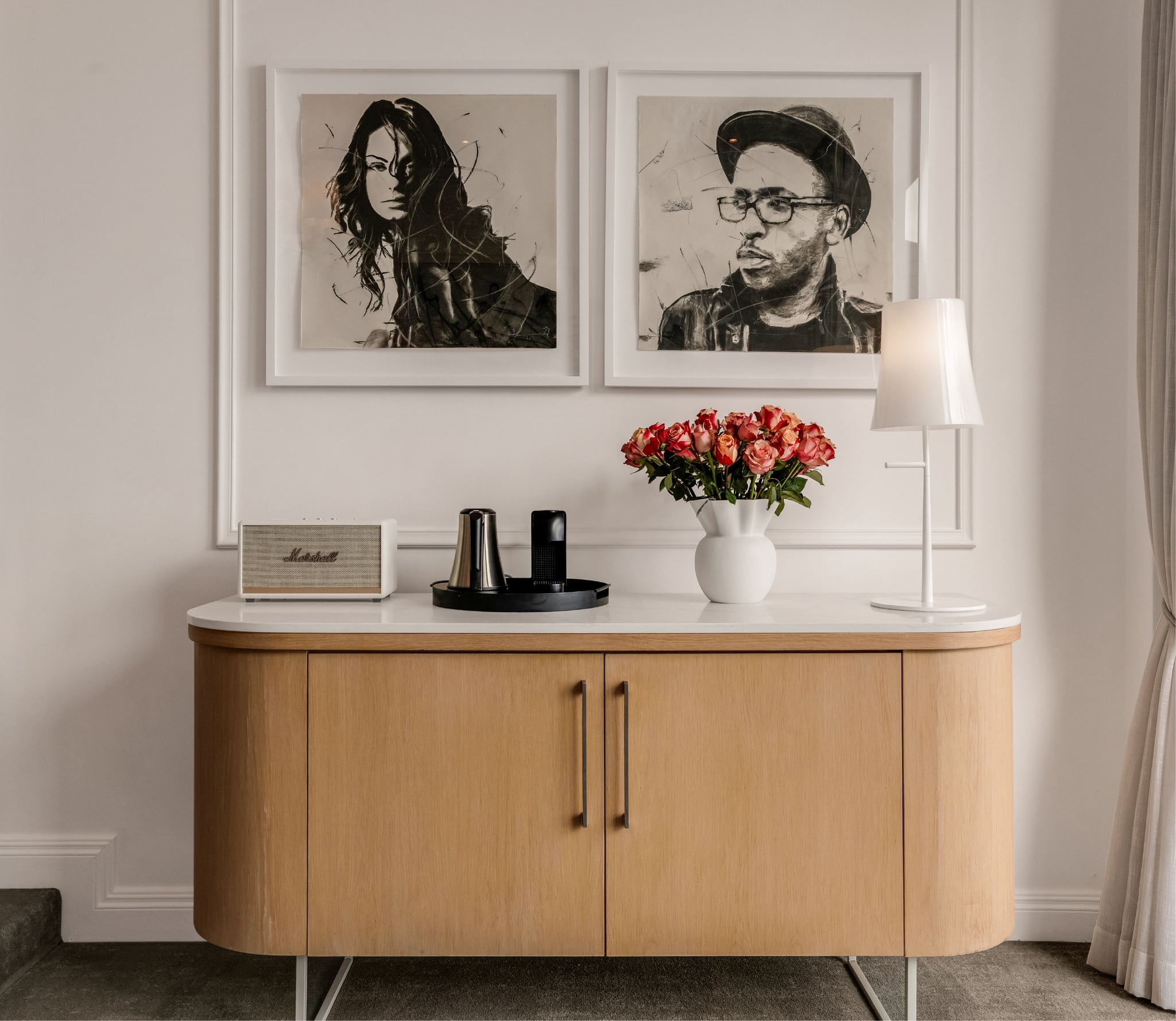 Charcoal portrait artworks above a wooden credenza with Marshall speaker, Nespresso machine and fresh roses in a Deluxe Sea-Facing Suite