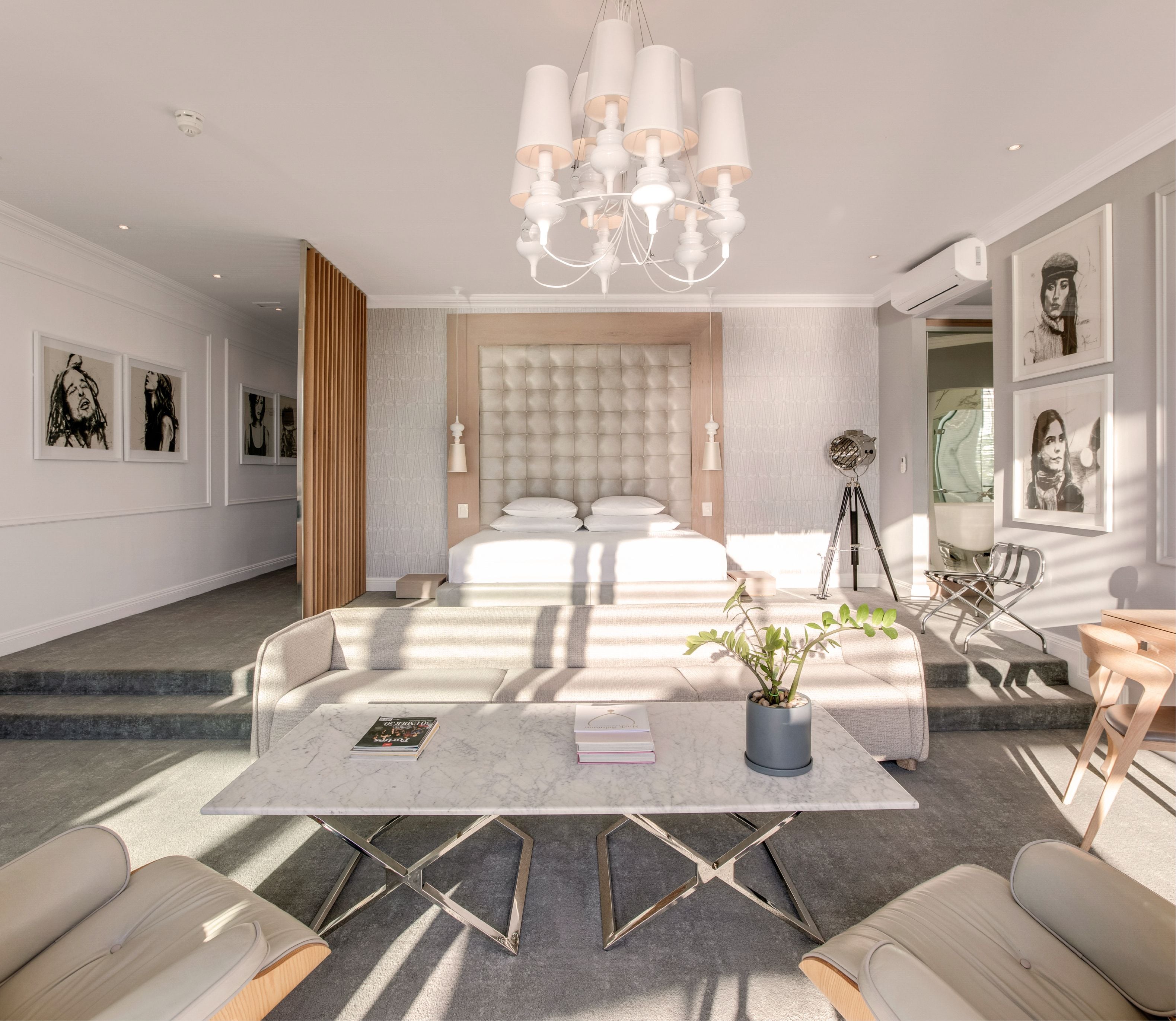 Wide-angle view of a luxury suite showing a tufted king bed framed by timber slat panelling, a white chandelier overhead, marble coffee table, cream sofas, and gallery walls of black-and-white portrait artworks on either side.