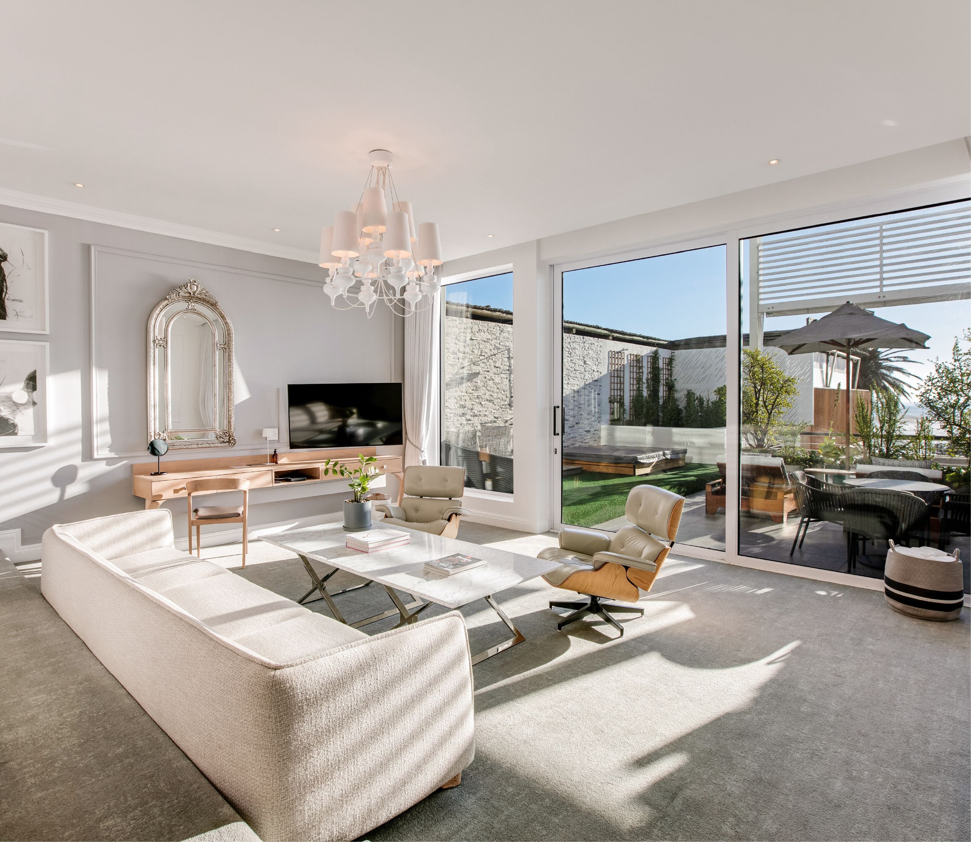 Spacious suite living area with a cream sofa, Eames lounge chairs, and marble coffee table bathed in natural light, with a gilt mirror, timber console, and large sliding glass doors opening onto a planted terrace with sea glimpses.