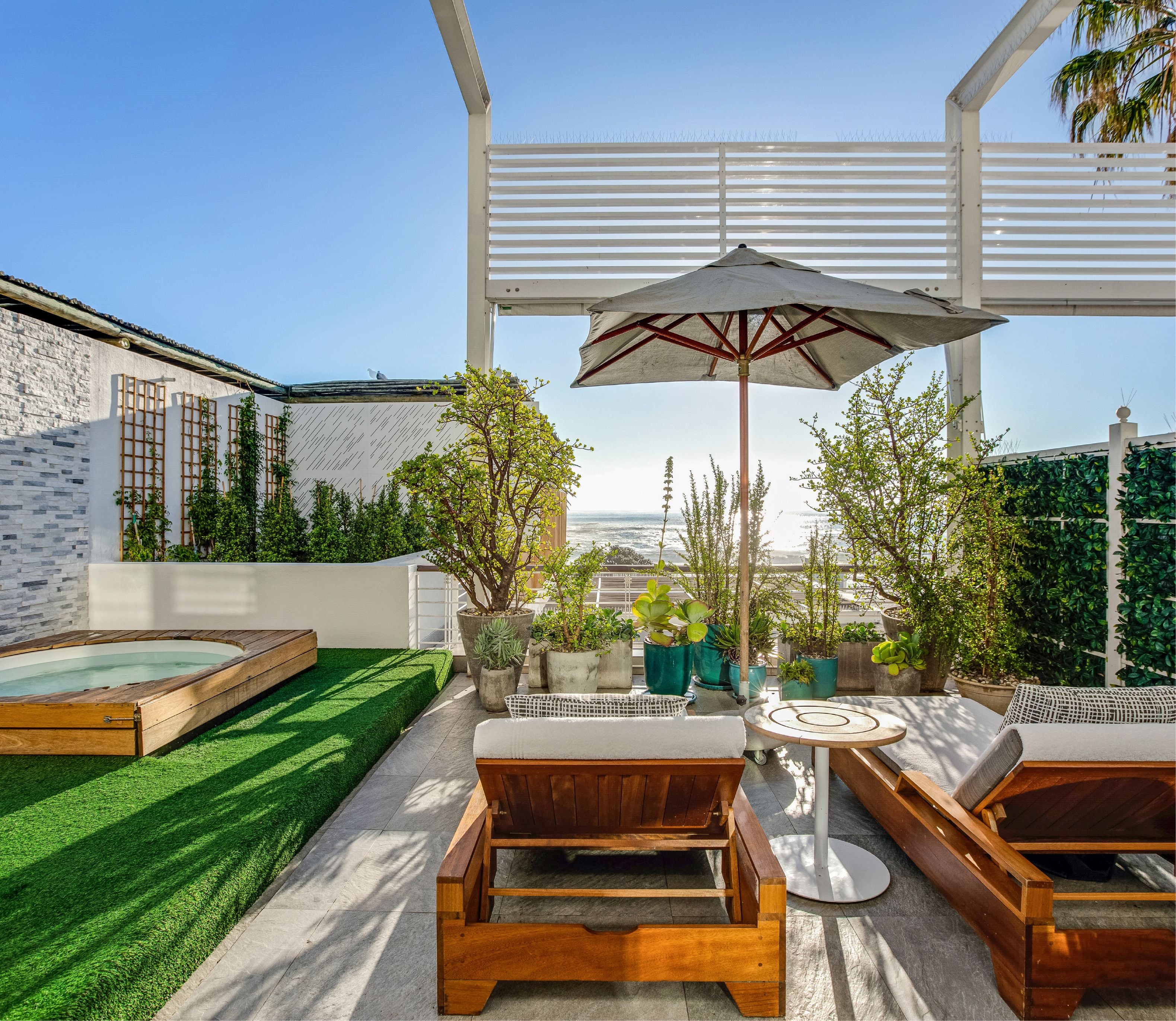 Rooftop terrace with a built-in jacuzzi set into a timber-decked lawn area, teak sun loungers, a round side table beneath a large umbrella, and lush potted greenery framing an open ocean view under a clear blue sky.