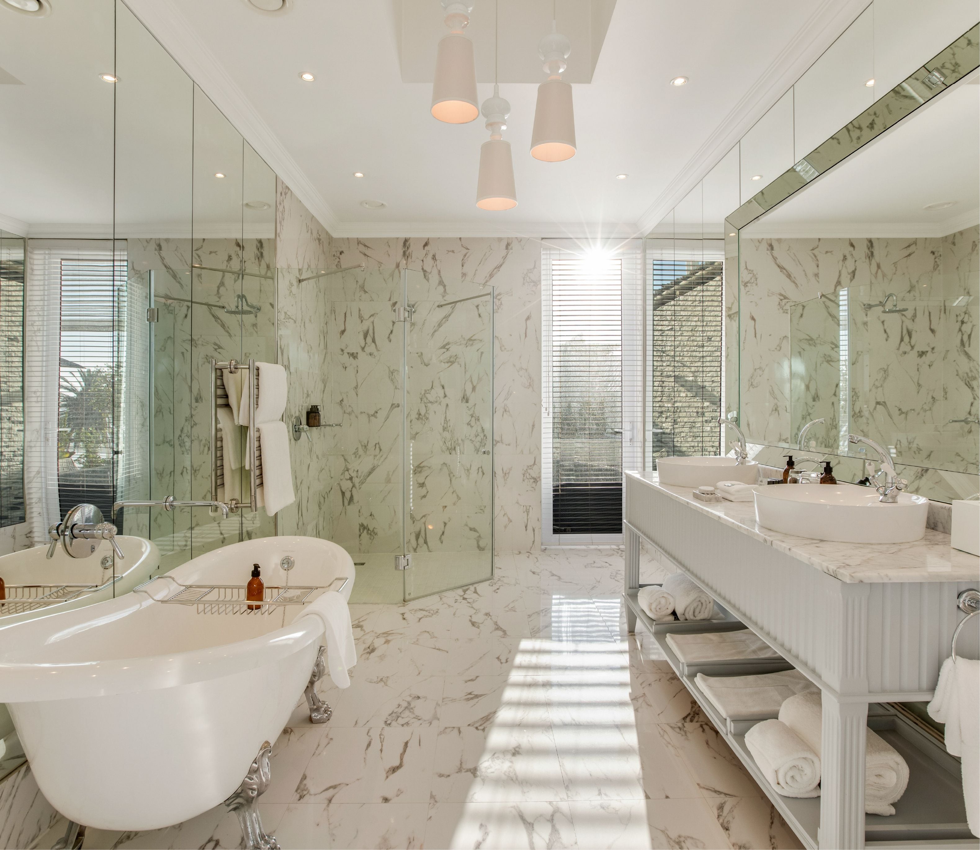 Sunlit luxury bathroom clad floor-to-ceiling in white and grey marble, featuring a freestanding clawfoot bathtub, glass-enclosed walk-in shower, double vessel sink vanity with stacked white towels, and pendant lighting overhead.