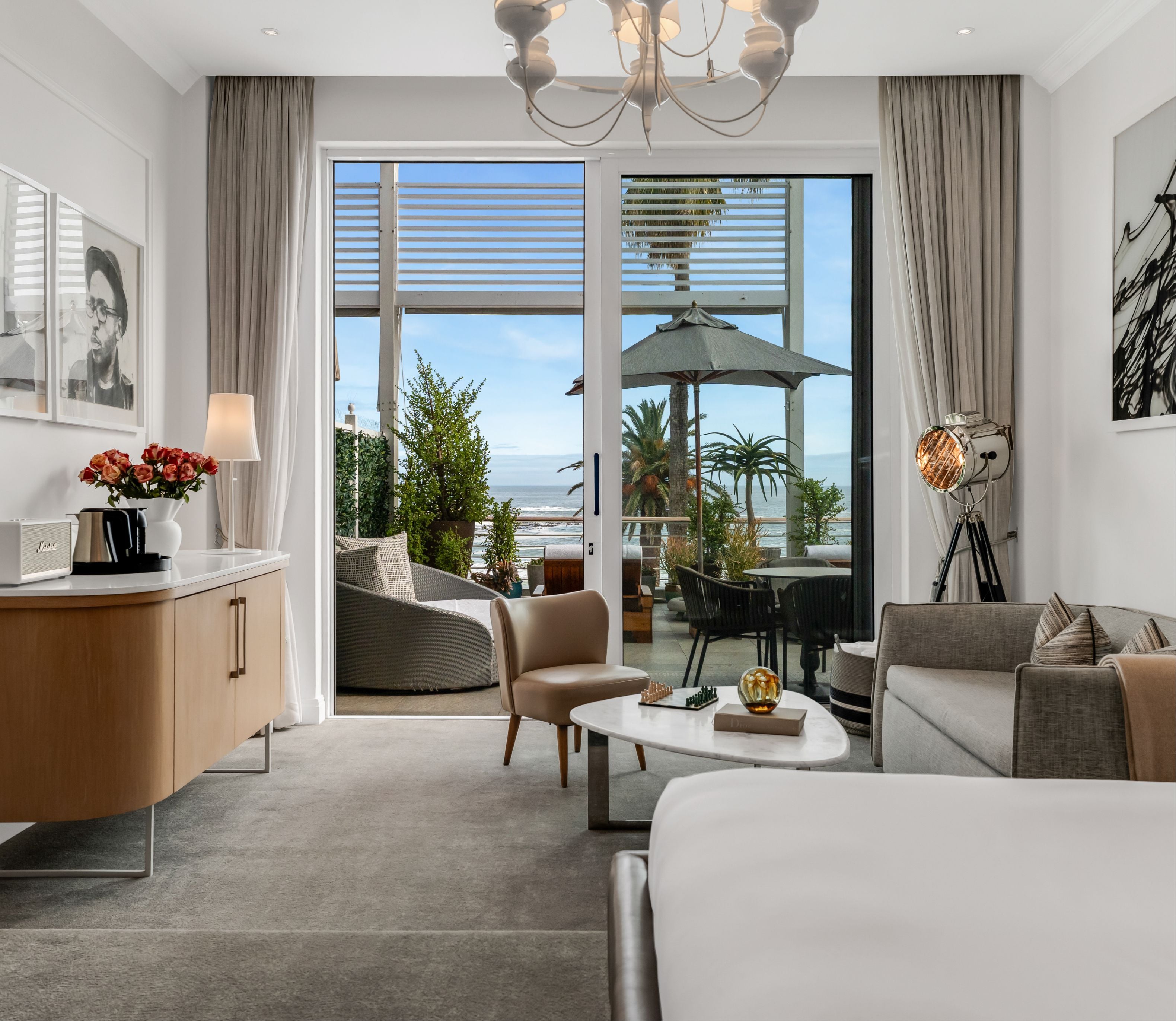 Bright suite interior with a king bed, warm-toned timber sideboard with fresh flowers and a Marshall speaker, and floor-to-ceiling glass doors opening to a furnished terrace with sea and palm tree views.