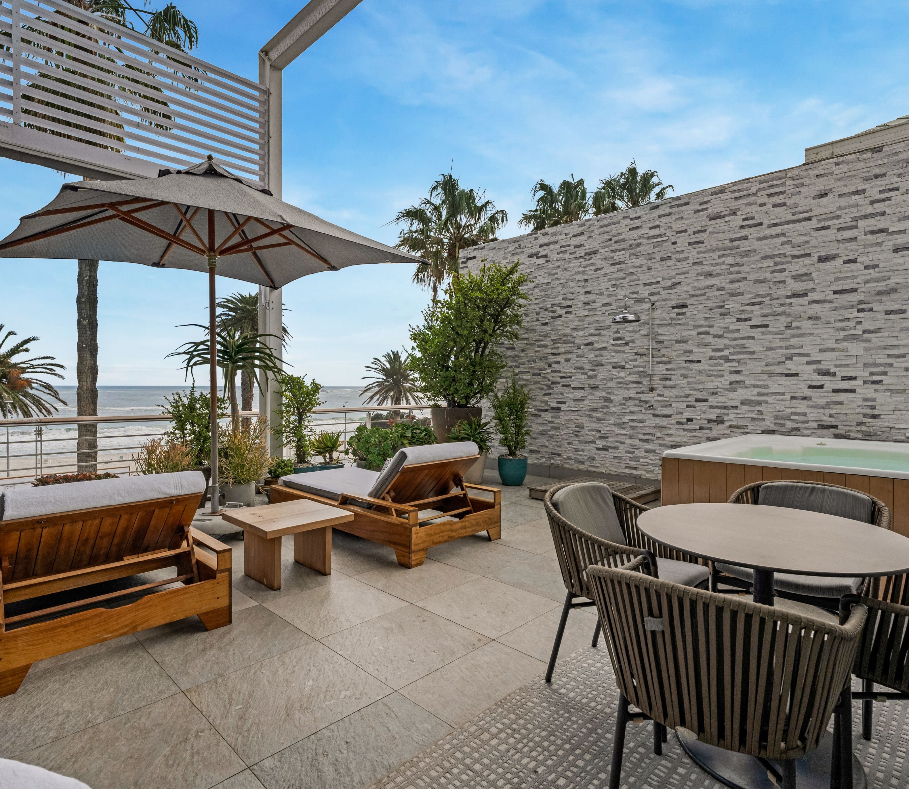 Expansive private terrace featuring teak sun loungers, an outdoor dining set, and a built-in jacuzzi beside a stone-clad wall, with palm trees and sea views in the background.