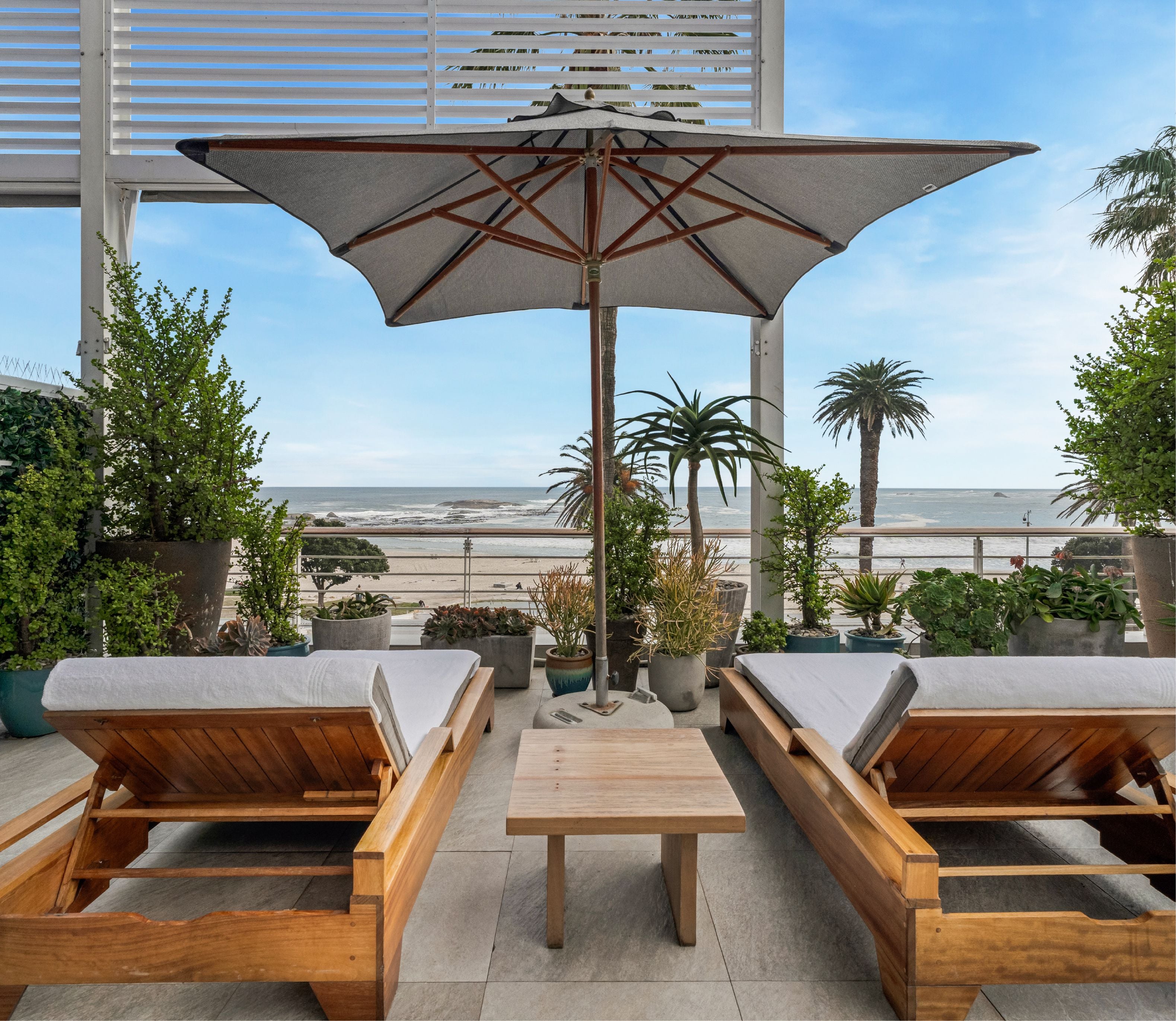 Private terrace with two teak sun loungers and a low coffee table beneath a large umbrella, surrounded by lush potted plants with an unobstructed view of the beach and ocean beyond.